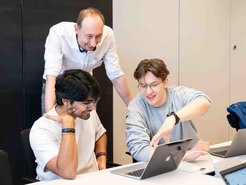 Carsten Rohr looks over the shoulders of Manoram Agarwal and Vincent Weis as they discuss a presentation slide.