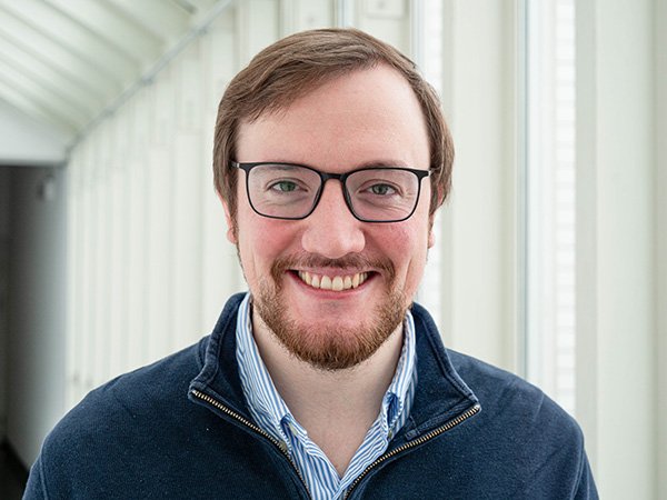 portrait photo of Maximilian Binzer: white male, brown hair and beard, glasses with a black frame, white and blue-striped shirt with dark blue pullover over it 